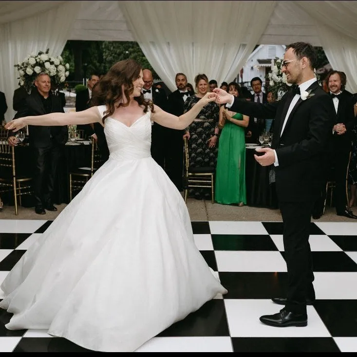 A bride and groom dance on a checkered floor surrounded by their guests